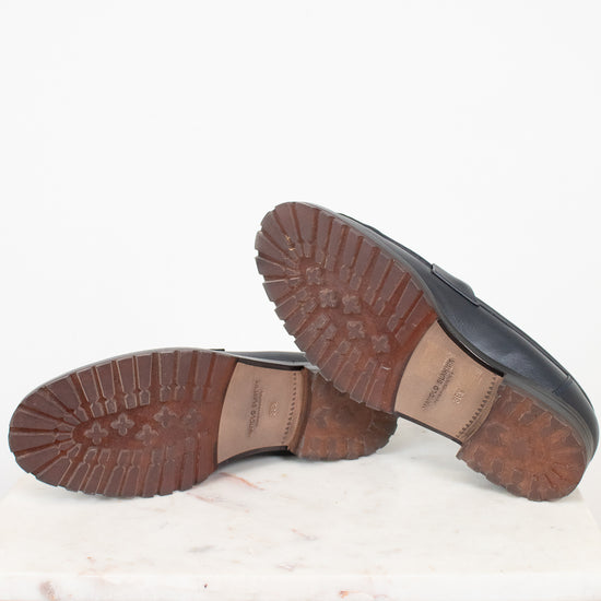 A pair of navy leather loafers shown from the bottom, displaying their brown lug soles, resting on a white marble slab against a white background.
