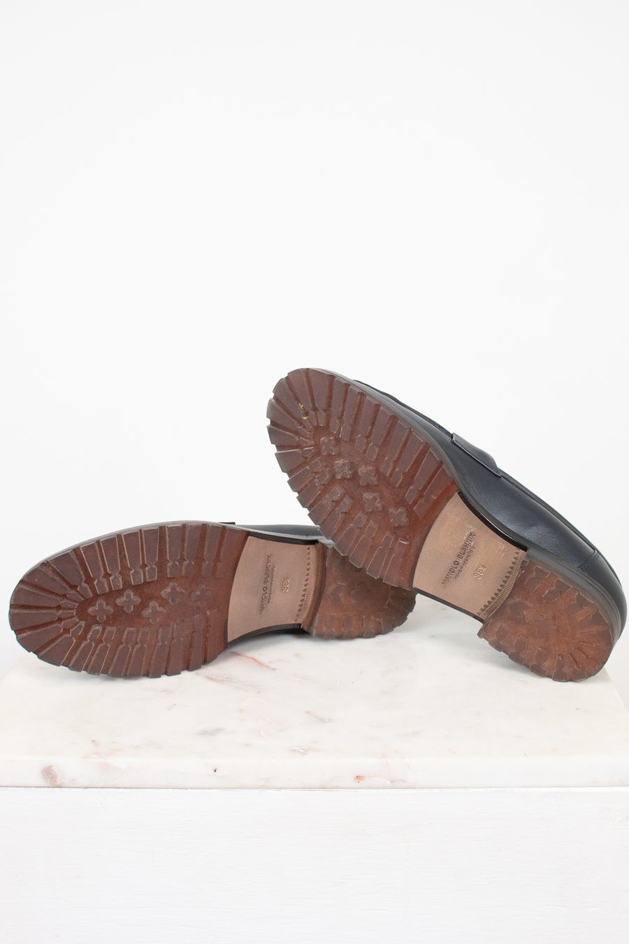 A pair of navy leather loafers shown from the bottom, displaying their brown lug soles, resting on a white marble slab against a white background.