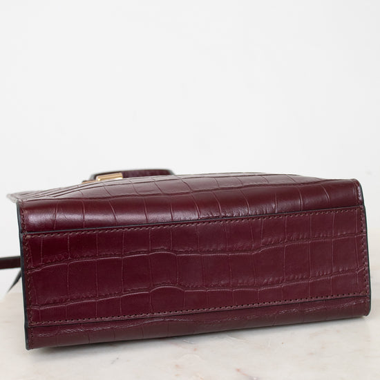 A dark burgundy crocodile-embossed leather clutch or wallet, shown from a side angle resting on a white marble surface against a white background.