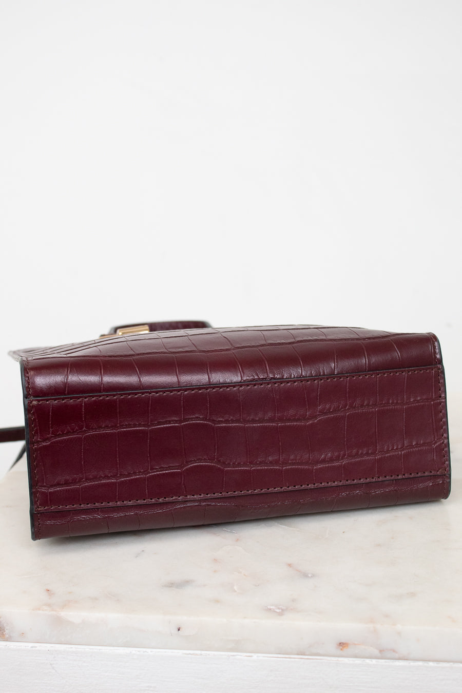 A dark burgundy crocodile-embossed leather clutch or wallet, shown from a side angle resting on a white marble surface against a white background.