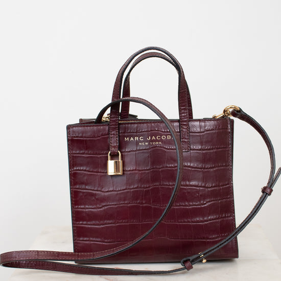A dark burgundy crocodile-embossed leather handbag by Marc Jacobs, featuring dual top handles, a detachable shoulder strap, a gold padlock, and gold logo lettering on the front, displayed on a light marble surface against a white background.