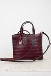 A dark burgundy crocodile-embossed leather handbag by Marc Jacobs, featuring dual top handles, a detachable shoulder strap, a gold padlock, and gold logo lettering on the front, displayed on a light marble surface against a white background.