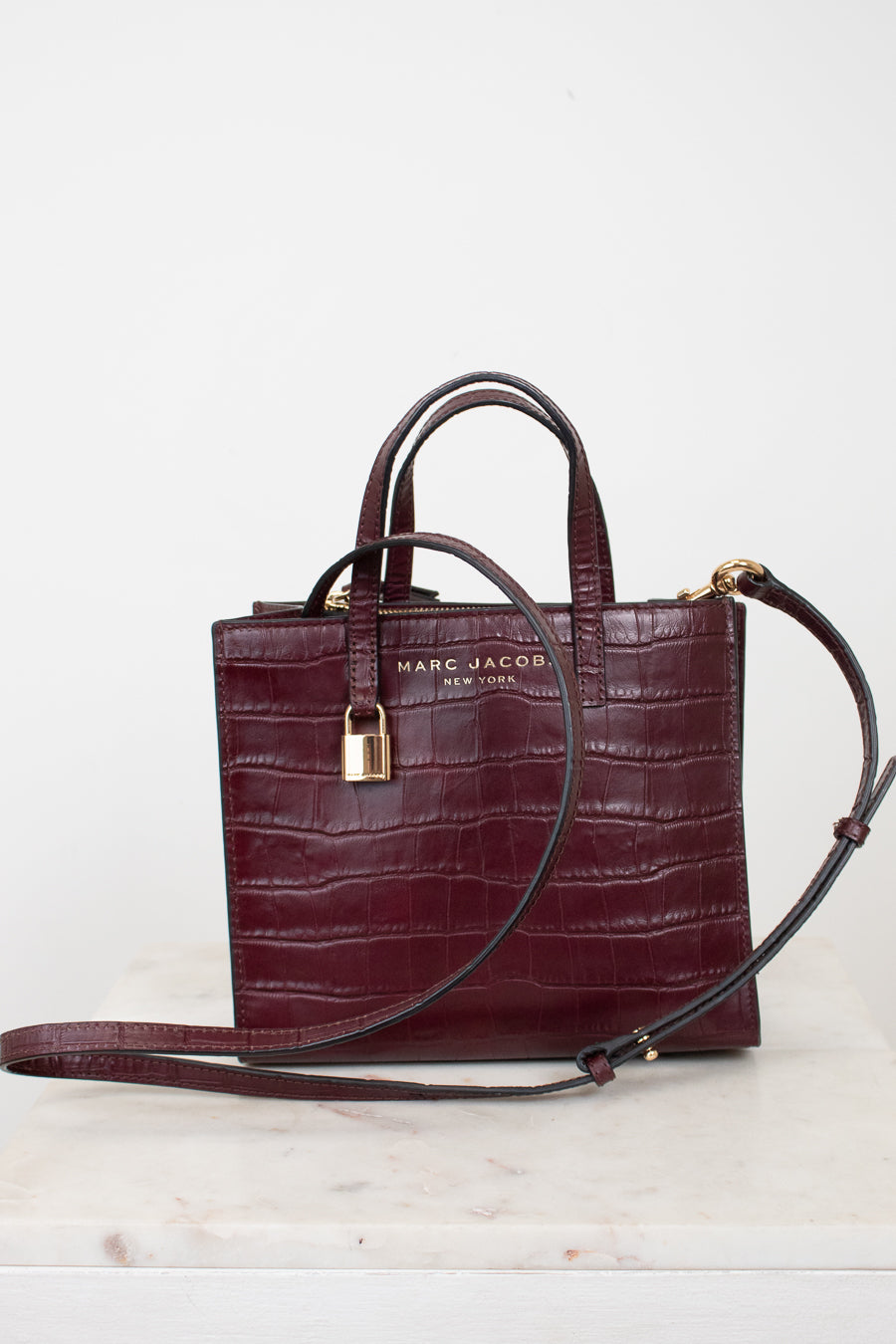 A dark burgundy crocodile-embossed leather handbag by Marc Jacobs, featuring dual top handles, a detachable shoulder strap, a gold padlock, and gold logo lettering on the front, displayed on a light marble surface against a white background.