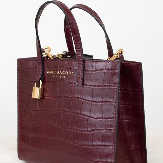 A Marc Jacobs New York crocodile-embossed leather tote bag in deep burgundy, featuring dual top handles, gold-tone hardware, and a gold brand plaque on the front, set against a clean white background.
