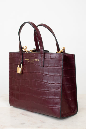 A Marc Jacobs New York crocodile-embossed leather tote bag in deep burgundy, featuring dual top handles, gold-tone hardware, and a gold brand plaque on the front, set against a clean white background.