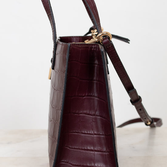 A structured dark burgundy crocodile-embossed leather handbag shown from a side profile view, resting on a light marble surface against a white background.