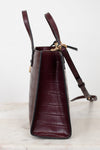 A structured dark burgundy crocodile-embossed leather handbag shown from a side profile view, resting on a light marble surface against a white background.