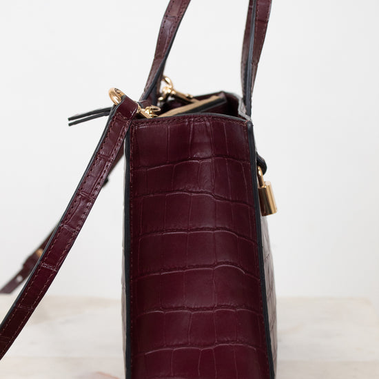 A structured handbag in dark burgundy crocodile-embossed leather, shown from the side profile on a light marble surface against a white background.