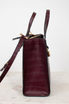 A structured handbag in dark burgundy crocodile-embossed leather, shown from the side profile on a light marble surface against a white background.