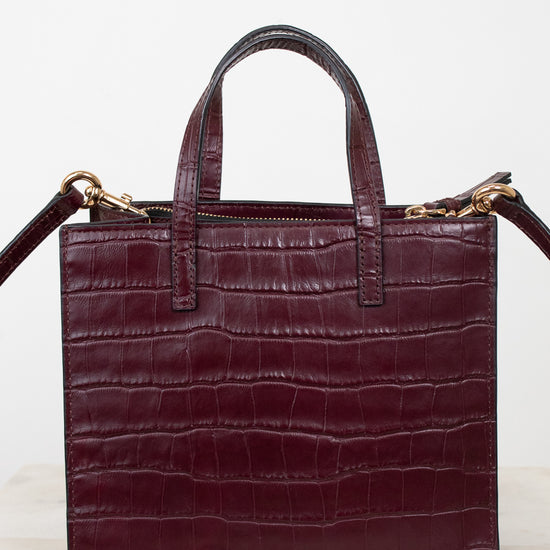 A structured burgundy crocodile-embossed leather handbag with double top handles and gold-tone hardware, displayed on a light marble surface against a white background.