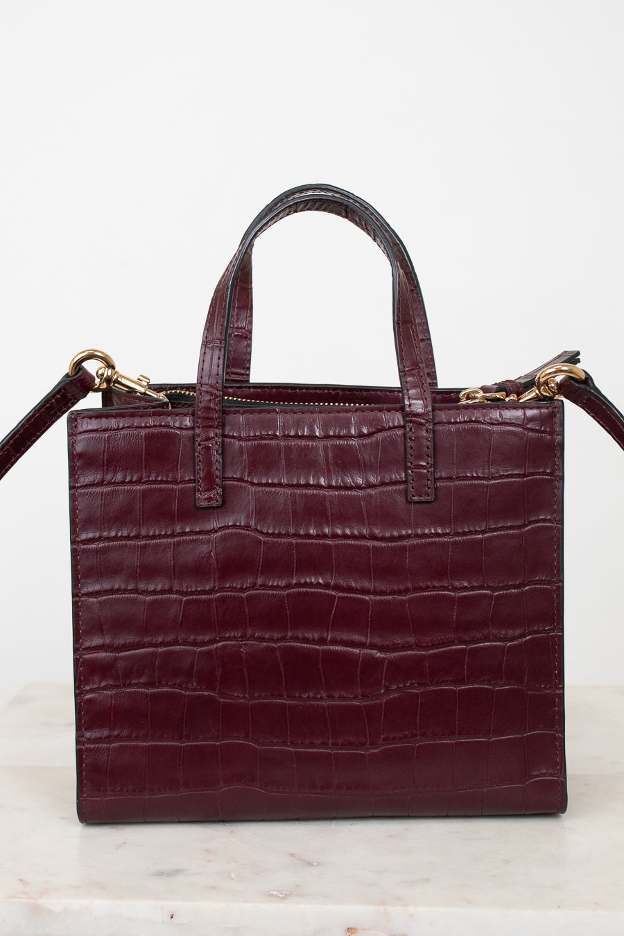 A structured burgundy crocodile-embossed leather handbag with double top handles and gold-tone hardware, displayed on a light marble surface against a white background.