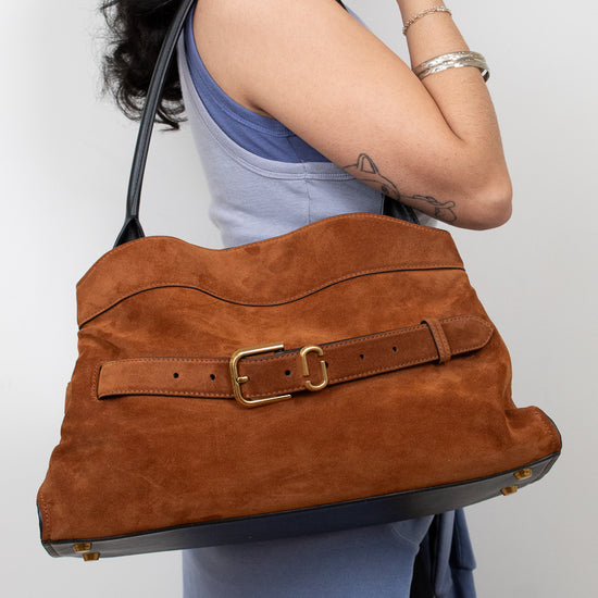 A woman wearing a light blue dress carries a large brown suede shoulder bag with a matching leather belt and gold buckle against a plain light gray background.