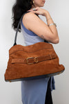 A woman wearing a light blue dress carries a large brown suede shoulder bag with a matching leather belt and gold buckle against a plain light gray background.