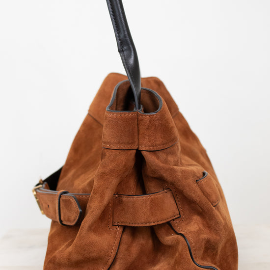 A brown suede bucket bag with black leather trim, a black shoulder strap, and a brown leather strap detail on the side, photographed against a plain white background.