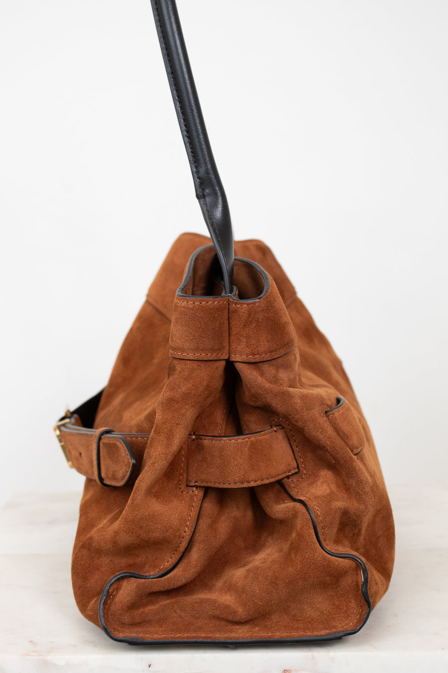 A brown suede bucket bag with black leather trim, a black shoulder strap, and a brown leather strap detail on the side, photographed against a plain white background.