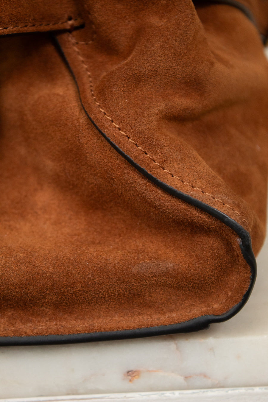 Close-up of a brown suede or nubuck bag corner with visible black piping along the edge, resting on a light marble surface.