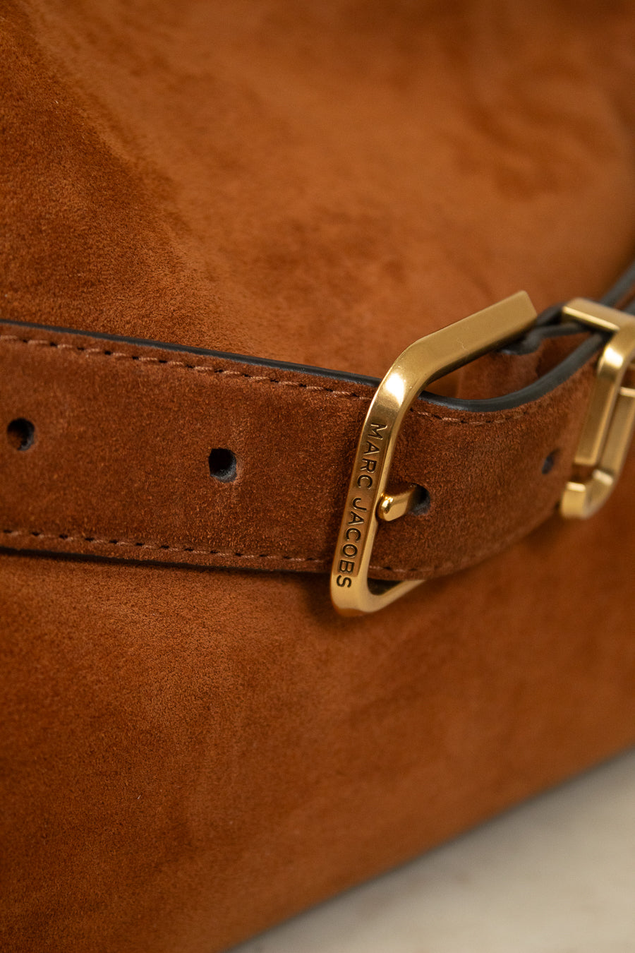 Close-up of a brown suede belt with a gold-tone Marc Jacobs buckle, attached to a matching brown leather strap on a brown suede bag.