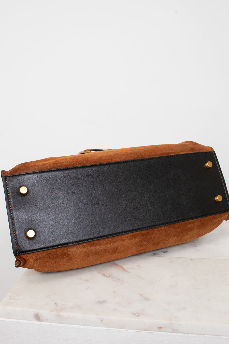 A close-up of the bottom corner of a brown suede and black leather handbag, showing gold-tone metal studs on a white marble surface against a white background.