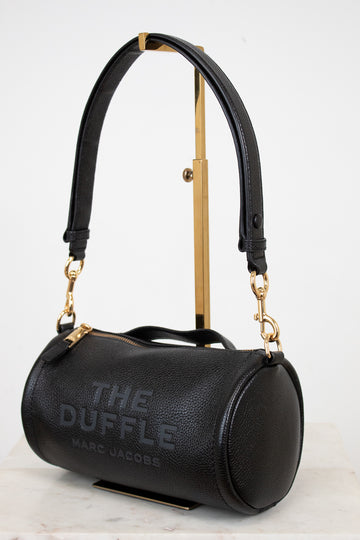 Black handbag with gold accents labeled 'The Duffle' by Marc Jacobs on a light background