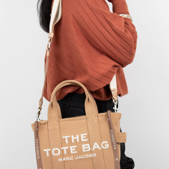 Person wearing a brown sweater and holding a tan tote bag with 'The Tote Bag' and 'Marc Jacobs' branding.