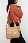 Person wearing a brown sweater and holding a tan tote bag with 'The Tote Bag' and 'Marc Jacobs' branding.