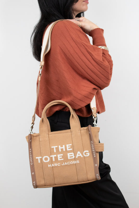 Person wearing a brown sweater and holding a tan tote bag with 'The Tote Bag' and 'Marc Jacobs' branding.