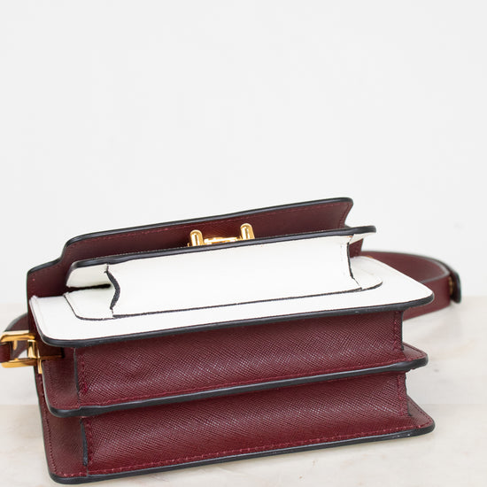 A structured leather handbag in deep burgundy saffiano with a white leather bottom panel and gold-tone hardware, displayed on a light marble surface against a white background.