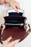 An open burgundy leather handbag with a dark navy blue interior lining and gold-tone hardware, held open by two hands wearing rings and bracelets against a plain white background.