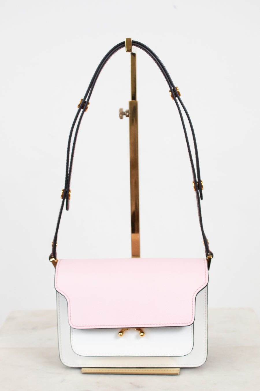 A structured shoulder bag with a light pink flap and a white body, featuring a black leather strap with gold-tone hardware, displayed on a minimalist stand.
