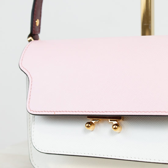 A close-up of a structured handbag with a light pink flap and a white lower body, accented by gold-tone hardware and a burgundy shoulder strap.