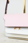A close-up of a structured handbag with a light pink flap and a white lower body, accented by gold-tone hardware and a burgundy shoulder strap.