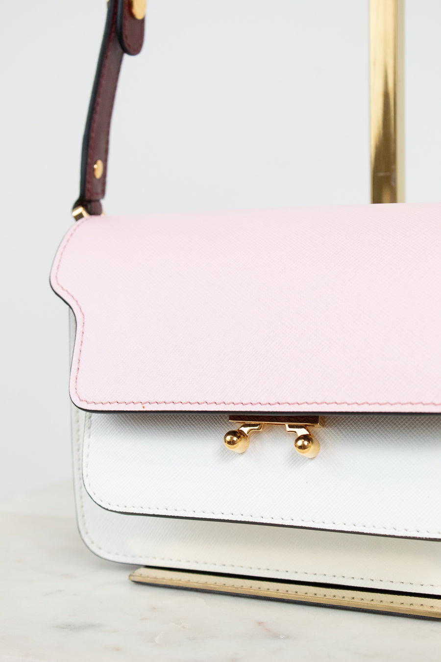 A close-up of a structured handbag with a light pink flap and a white lower body, accented by gold-tone hardware and a burgundy shoulder strap.