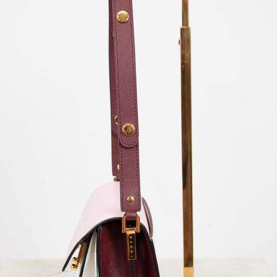 A structured leather handbag with a deep burgundy side panel and strap, with color-block details in pale blush pink, displayed on a gold metal stand against a clean white background.
