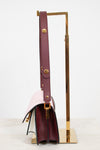 A structured leather handbag with a deep burgundy side panel and strap, with color-block details in pale blush pink, displayed on a gold metal stand against a clean white background.