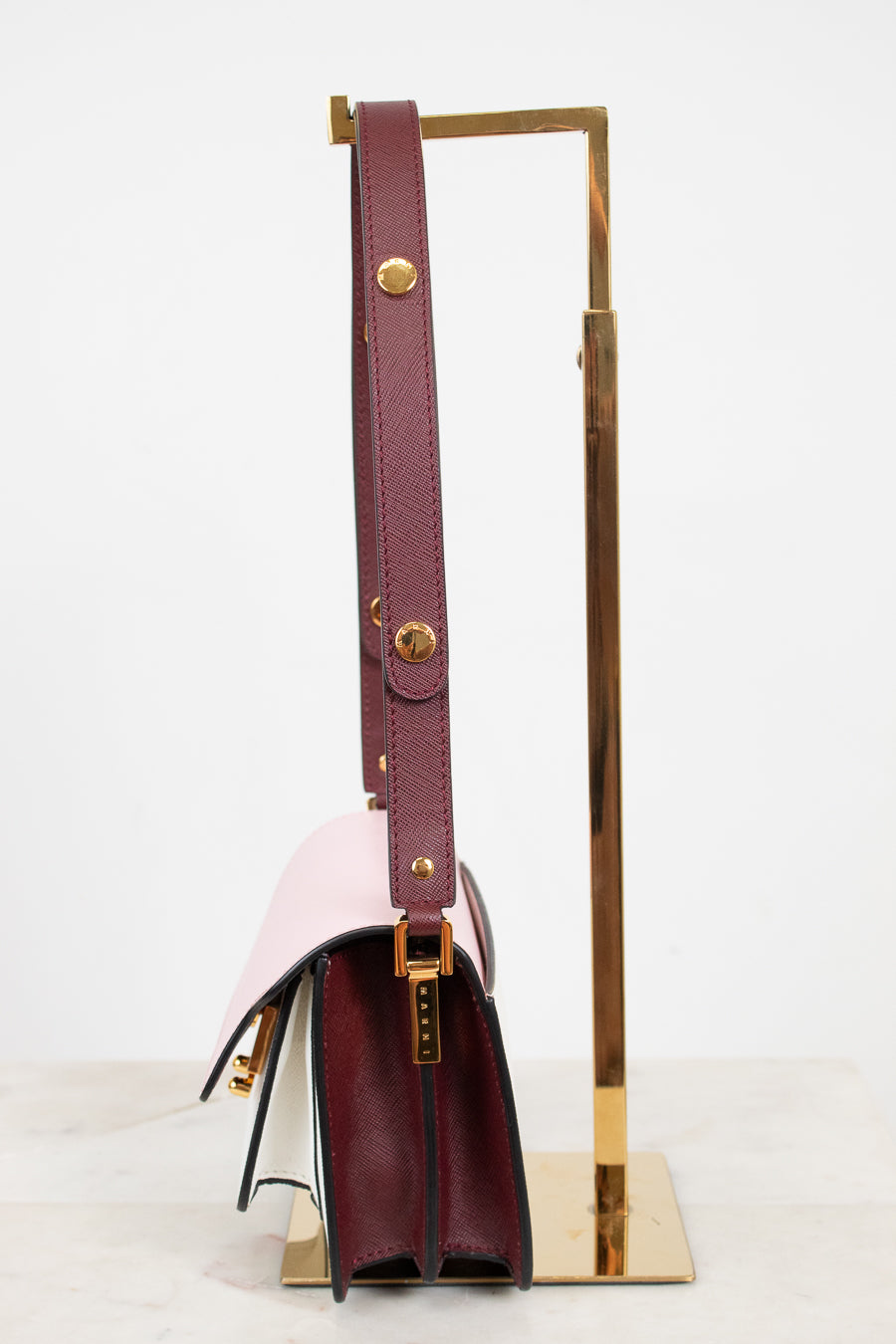 A structured leather handbag with a deep burgundy side panel and strap, with color-block details in pale blush pink, displayed on a gold metal stand against a clean white background.