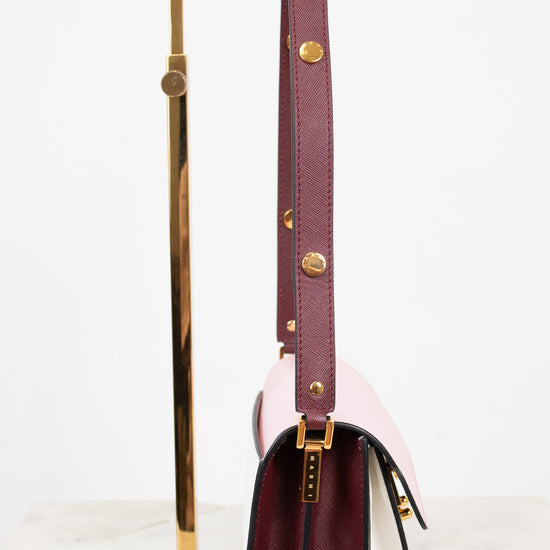 A structured leather shoulder bag in deep burgundy and pale blush pink, displayed on a gold metal stand against a clean white background.