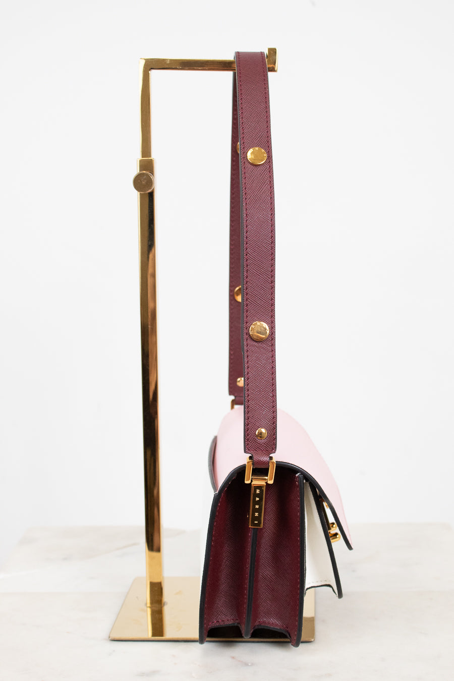 A structured leather shoulder bag in deep burgundy and pale blush pink, displayed on a gold metal stand against a clean white background.