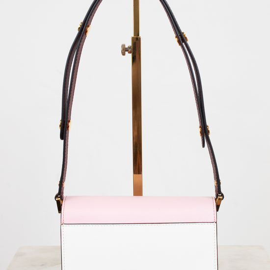 A structured rectangular handbag with a light pink top flap and white body, featuring burgundy leather shoulder straps with gold-tone hardware, displayed on a minimalist stand.