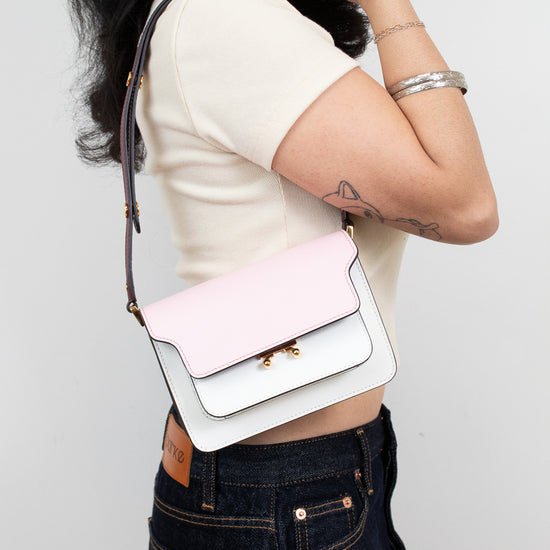 A woman wearing a cream short-sleeve crop top and dark blue jeans carries a small rectangular color-block bag with a pale pink flap and white body, featuring gold-tone hardware and a dark strap.
