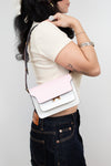A woman wearing a cream short-sleeve crop top and dark blue jeans carries a small rectangular color-block bag with a pale pink flap and white body, featuring gold-tone hardware and a dark strap.