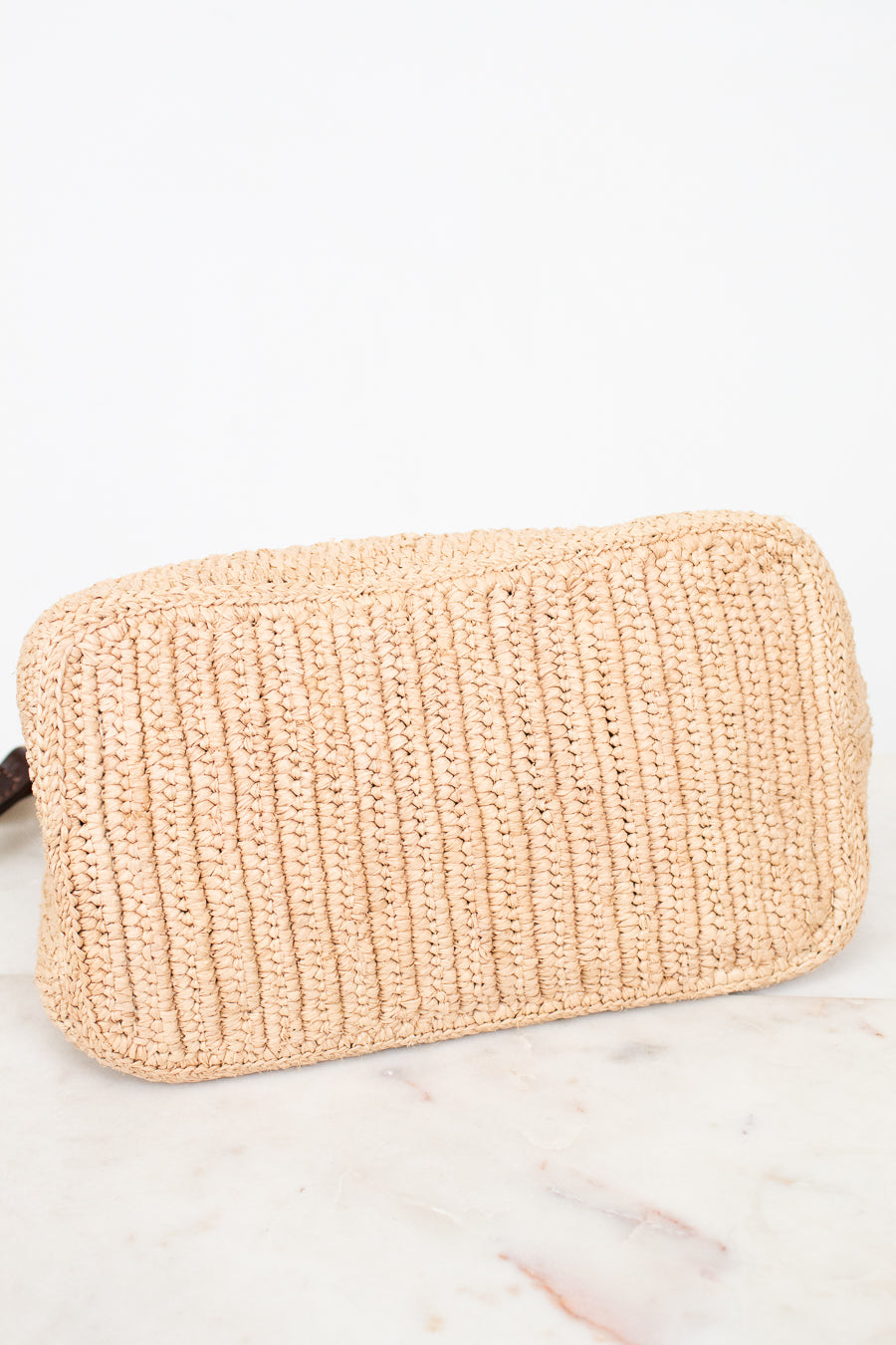 A light beige woven straw pouch with a ribbed texture, resting on a white marble surface against a white background.