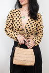 A woman wearing a tan and black polka dot long-sleeve tie-front top, black pants, and holding a woven straw handbag against a plain light background.