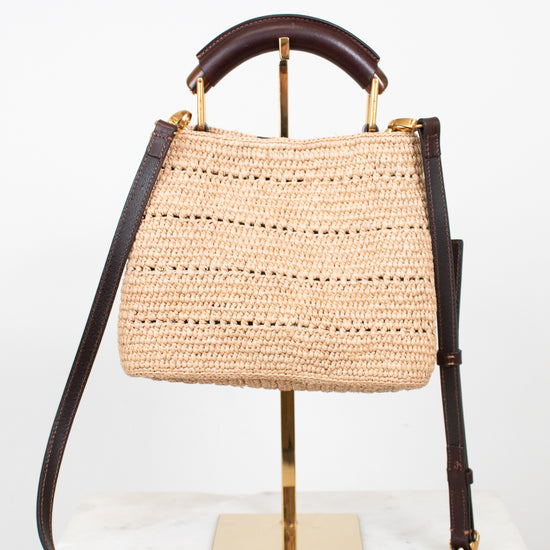A woven straw handbag with dark brown leather trim and a top handle, displayed on a gold stand against a white background.