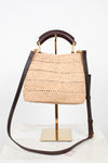 A woven straw handbag with dark brown leather trim and a top handle, displayed on a gold stand against a white background.