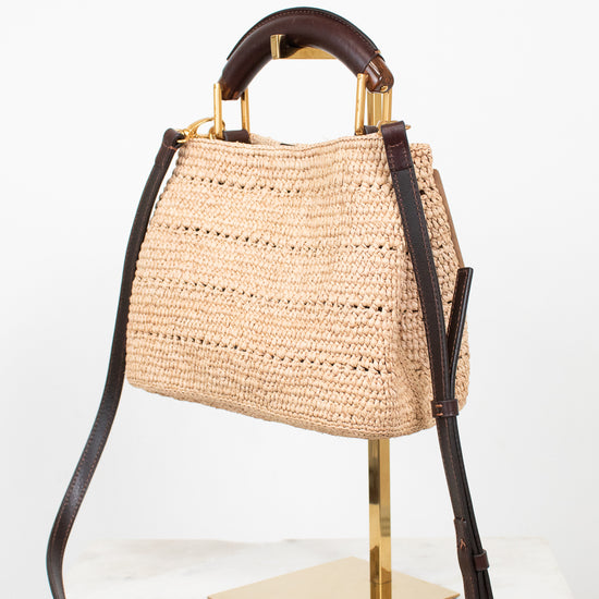 A woven straw handbag with dark brown leather top handle and shoulder strap, accented with gold-tone hardware, displayed on a gold stand against a white background.