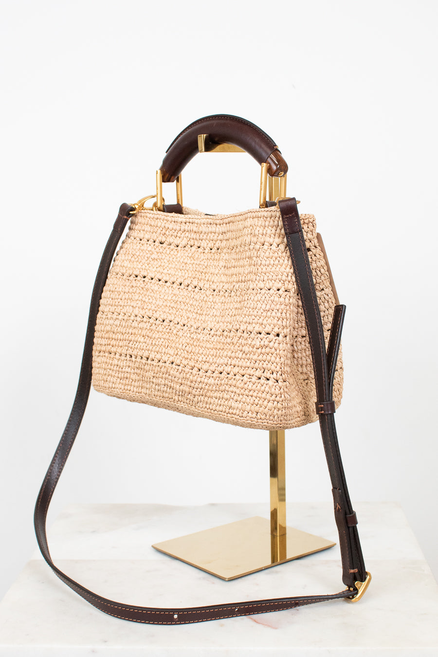 A woven straw handbag with dark brown leather top handle and shoulder strap, accented with gold-tone hardware, displayed on a gold stand against a white background.