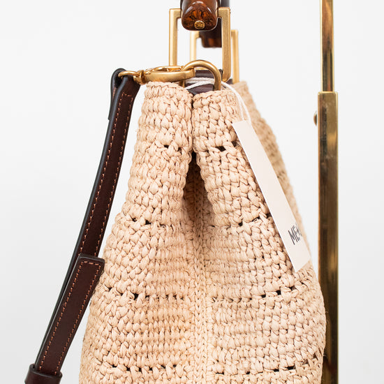 A woven straw bag in a light beige color with dark brown leather trim and strap, hanging from a gold metal stand against a white background.