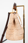 A woven straw bag in a light beige color with dark brown leather trim and strap, hanging from a gold metal stand against a white background.