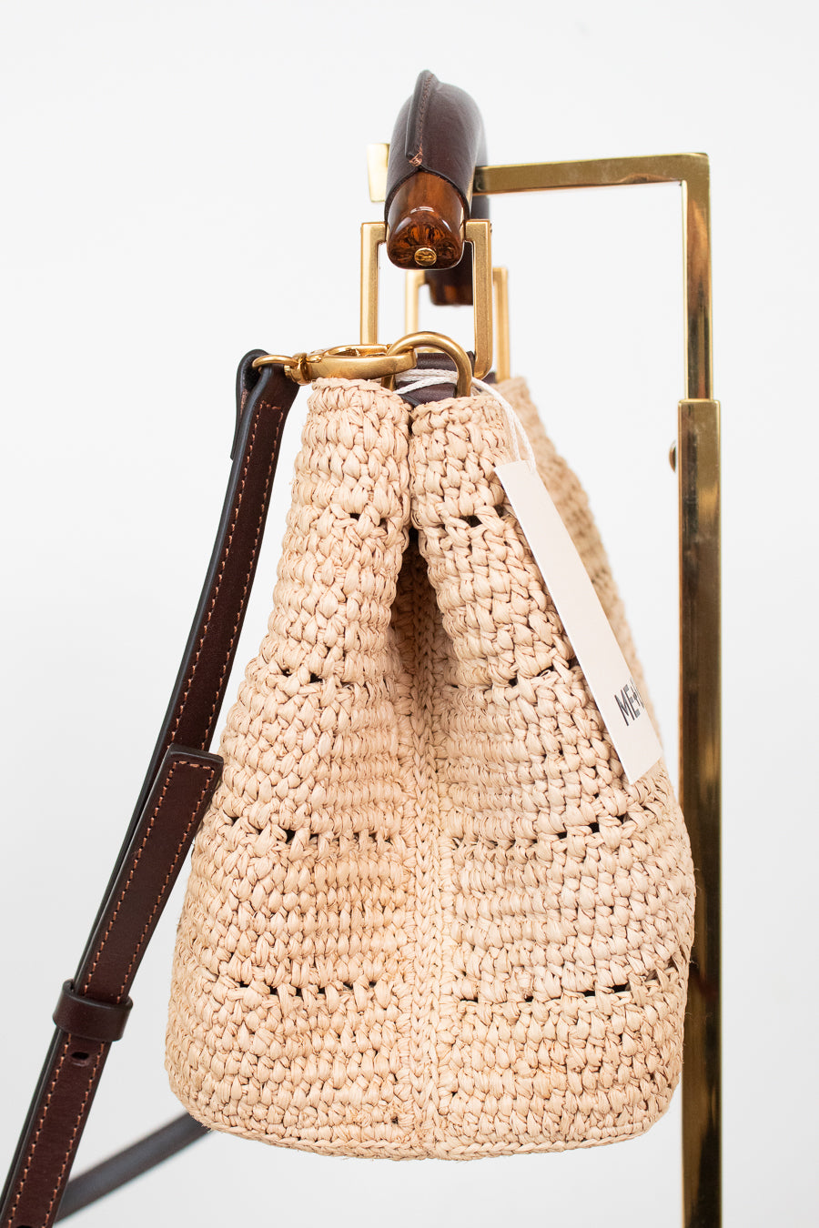 A woven straw bag in a light beige color with dark brown leather trim and strap, hanging from a gold metal stand against a white background.