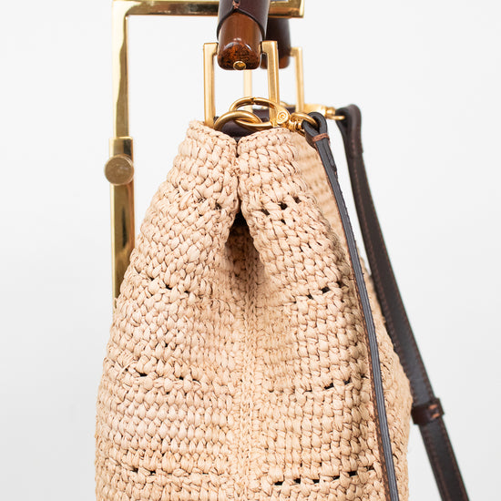 A natural beige woven straw bucket bag with a dark brown leather strap and gold-tone hardware, hanging on a gold metal display stand against a white background.